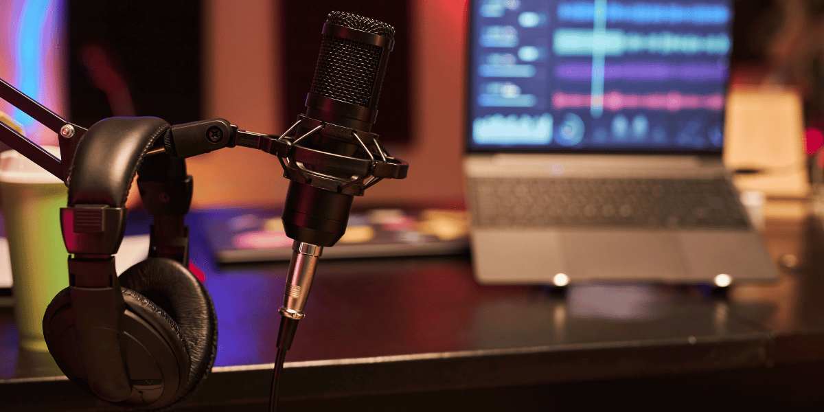 Podcast microphone and headphones beside a laptop with audio editing software on screen.