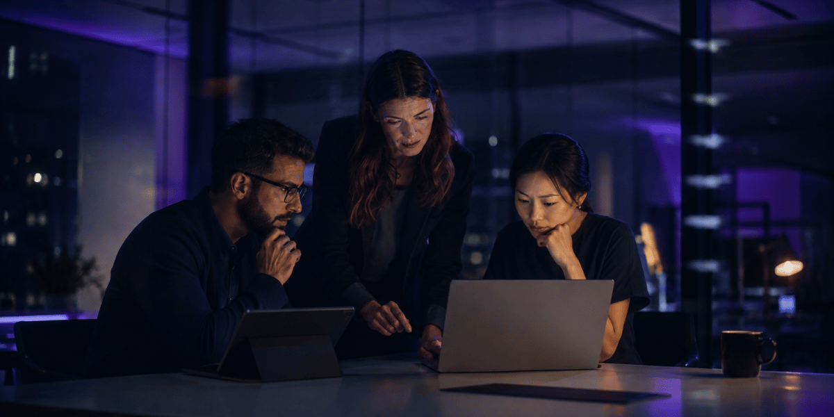 Three professionals review a laptop together in a dark, modern office with blue and purple lighting, representing HubSpot Credits strategy and AI usage planning.